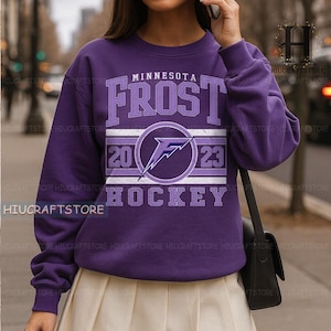 May include: A purple long-sleeve sweatshirt featuring the text Minnesota Frost Hockey in light purple lettering. The center graphic includes a stylized letter F inside a circle, flanked by the numbers 20 and 23, all set against a horizontal striped background.