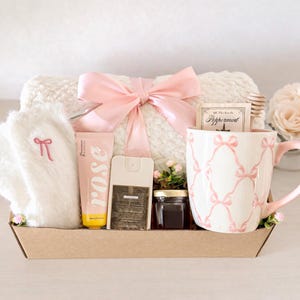 May include: A gift basket with a white blanket tied with a pink bow, a pink rose-scented lotion, a mug with a pink bow pattern, a small jar of honey, and a bottle of Velvet Vanilla spray. The basket also includes a pair of white fuzzy socks with a pink bow.
