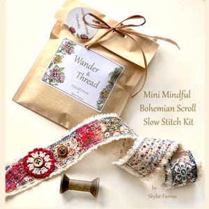 Può includere: Un kit artigianale con un pacco di carta marrone legato con un nastro, un rotolo di tessuto decorativo e una bobina di legno. Il rotolo presenta un tessuto fantasia rosso, bianco e blu con un fiore e un bottone. Il testo sulla confezione recita "Wander & Thread".