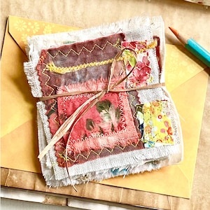 Peut inclure: Un ensemble de carrés de tissu avec des motifs floraux et imprimés, cousus avec du fil jaune et rouge. L'ensemble est noué avec un nœud en raphia et repose sur une enveloppe jaune, avec un crayon en arrière-plan.