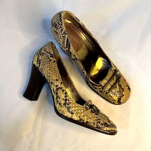 Könnte beinhalten: Ein Paar hochhackige Pumps mit Schlangenledermuster in Beige-, Braun- und Schwarztönen. Die Schuhe haben eine eckige Schuhspitze, einen dunkelbraunen Blockabsatz und ein dekoratives Schnallendetail auf der Vorderseite. Das Innenfutter ist goldfarben.