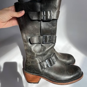 Può includere: Stivali neri in pelle con tacco e più fibbie. Gli stivali hanno un tacco in legno marrone.