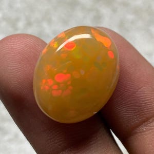 May include: A polished, oval-shaped opal gemstone. The opal has a warm, yellow-orange base color with vibrant red and orange internal fire patterns. The gemstone is held between fingers, showcasing its size and luster. This gemstone is suitable for jewelry making.