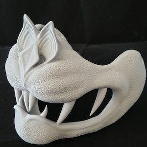 Puede incluir: Una máscara blanca impresa en 3D de un monstruo de dibujos animados con dientes grandes y orejas puntiagudas. La máscara está hecha de plástico texturizado y tiene un agujero para los ojos.