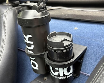 May include: Black shaker bottle and travel mug in a car cup holder. The shaker has white text that says "bouk" and the travel mug has the text "CIO industrial design". The cup holder is black and mounted on a blue surface.