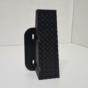Pode incluir: Degrau de plástico preto com um padrão de banda de rodagem de placa de diamante. O degrau tem um suporte de montagem lateral.