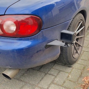 May include: A blue car's rear with a GoPro camera mounted on a black bracket near the tire. The car has a red tail light and a silver exhaust pipe. The tire is black.