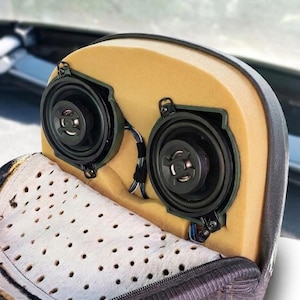 Op de afbeelding: Een hoofdsteun voor in de auto met de buitenste stof gedeeltelijk opengeritst, waardoor twee ronde zwarte luidsprekers in een gele schuimkern zichtbaar zijn. De luidsprekers zijn verbonden met zichtbare draden en een geperforeerde witte voering.