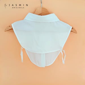 May include: A white detachable collar with a classic design, hanging on a wooden hanger. The collar has a pointed collar and ties at the front. The brand name "JASMIN ORIGINALS" is visible at the top left.