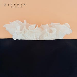 Pode incluir: Uma peça de roupa preta com acabamento branco franzido sobre um fundo pêssego. A parte superior da peça apresenta um tecido delicado e plissado que adiciona textura e interesse visual. A marca "JASMIN ORIGINALS" está impressa no topo.