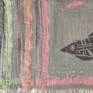 Puede incluir: Una bufanda gris con rayas verticales en verde, melocotón y rosa. Un diseño de pez está en el lado derecho. La bufanda tiene un borde con flecos y el texto "JASMIN ORIGINALS".