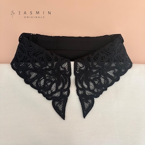 May include: A black detachable collar featuring an intricate floral lace pattern with scalloped edges. The text JASMIN ORIGINALS is printed in the upper left corner. The collar is displayed against a light-coloured fabric background.