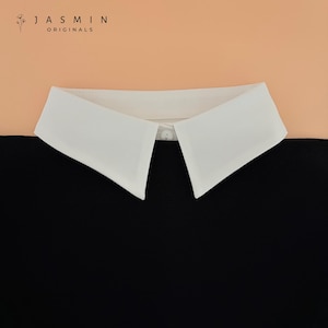 Op de afbeelding: Een witte afneembare kraag met een knoopsluiting, tegen een perzik- en zwarte achtergrond. De kraag is scherp en hoekig, met een klassiek puntig ontwerp. De merknaam "JASMIN ORIGINALS" staat linksboven gedrukt.