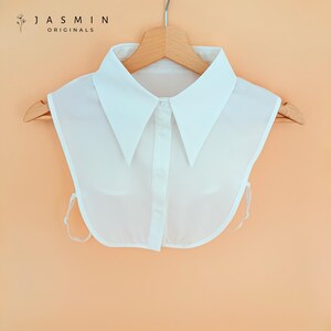 May include: A white detachable collar with a pointed collar design, button-down front, and tie closures. The collar is hanging on a wooden hanger. The text "JASMIN ORIGINALS" is printed at the top left.
