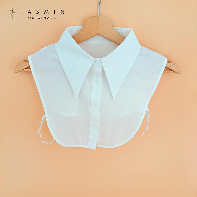 May include: A white detachable collar with a pointed collar design, button-down front, and tie closures. The collar is hanging on a wooden hanger. The text "JASMIN ORIGINALS" is printed at the top left.