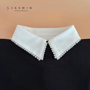 Puede incluir: Un cuello blanco desmontable con diseño en punta y un borde de pequeñas perlas blancas redondas a lo largo de los extremos. El texto JASMIN ORIGINALS es visible en la esquina superior izquierda sobre el cuello, colocado sobre un fondo negro.