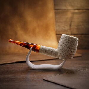 May include: A tobacco pipe featuring a textured, off-white bowl with a repeating circular pattern and a smooth, translucent amber-colored stem. The pipe is supported by a curved, white stand that cradles the stem and the base of the bowl.