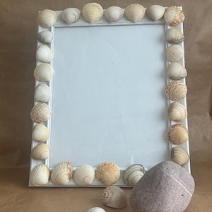 May include: White picture frame decorated with seashells. The frame is sitting on a brown surface with a small grey rock and a seashell.