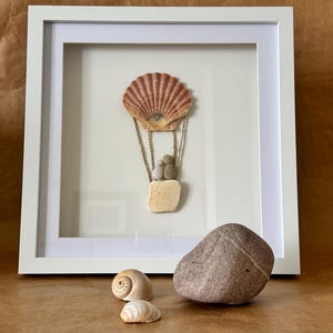 May include: A white framed picture featuring a seashell hot air balloon crafted from natural materials. The balloon is made from a large pink seashell with a smaller seashell basket and stones as passengers. The picture is on a brown surface with two smaller seashells and a large grey rock.