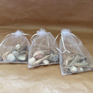 May include: Three white organza bags filled with seashells, driftwood, and twine. The bags are tied with white ribbons and sit on a brown paper surface. The shells vary in size and colour, with shades of white, tan, and brown.