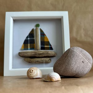 May include: A framed artwork featuring a sailboat made from driftwood and fabric. The sailboat has a yellow, black, and green plaid sail. The artwork is displayed on a table with a brown stone and two seashells.