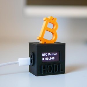 May include: A small black box with a screen displaying "BTC Price: $ 98.048" and the word "HODL". A 3D printed orange Bitcoin symbol sits on top of the box.