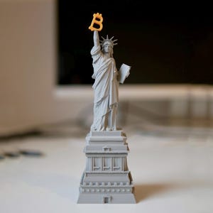 May include: A gray 3D printed model of the Statue of Liberty holding a yellow plastic letter "B" above her head. The statue is standing on a gray base with a detailed design.
