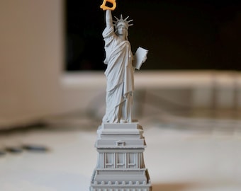 May include: A gray 3D printed model of the Statue of Liberty holding a yellow plastic letter "B" above her head. The statue is standing on a gray base with a detailed design.