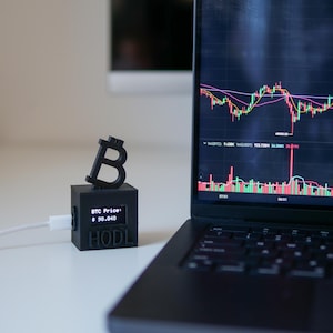 Peut inclure: Un ordinateur portable noir avec un écran affichant un graphique de crypto-monnaie. Le graphique montre un graphique en ligne rouge et vert avec une ligne rose. L'ordinateur portable est sur une surface blanche à côté d'un symbole Bitcoin imprimé en 3D noir avec un petit écran qui affiche "BTC Price: $ 98.048 HODL".