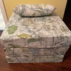 May include: A stack of three cushions with a map-like pattern in shades of green, blue, and white. The top cushion has a matching pillow on top.