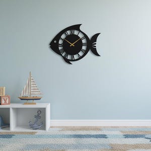 May include: A black fish-shaped wall clock with Roman numerals and gold hands hangs on a light blue wall. A white shelf below holds nautical-themed decor, including a sailboat and a whale.