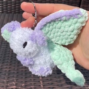 May include: A handmade, plush moth keychain in white, lavender, and mint green. The moth has a fluffy body, lavender accents, and mint green wings. A silver-colored metal clasp is attached for easy use.