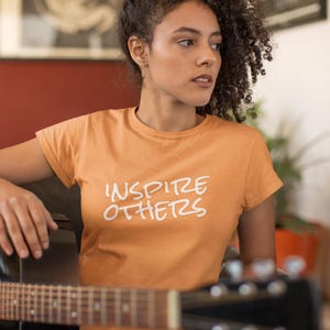 May include: A woman wearing an orange t-shirt with the text "Inspire Others" in white lettering. She is holding an acoustic guitar.