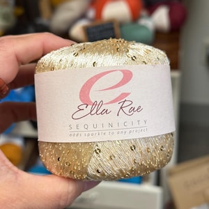 May include: A gold yarn ball with sequins, labeled "Ella Rae SEQUINICITY adds sparkle to any project." The yarn is held in a hand, with other yarn balls in the background. This yarn is ideal for knitting and crochet projects.