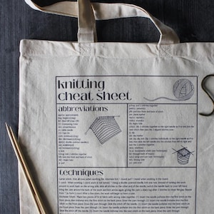 Puede incluir: Bolso tote color crema con un gráfico en blanco y negro de "Knitting Cheat Sheet". El bolso presenta abreviaturas y técnicas de tejido, junto con dos agujas de tejer de madera.
