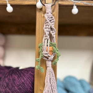 May include: A small macrame plant hanger with a terracotta pot and green faux plants. The hanger is made with a light pink cord and has a tassel at the bottom.