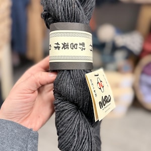 May include: A skein of dark charcoal gray yarn with a textured, slightly heathered appearance. It features a wide paper band with Japanese text and a smaller, cream-colored tag displaying the Noro logo and the text The World of Nature.