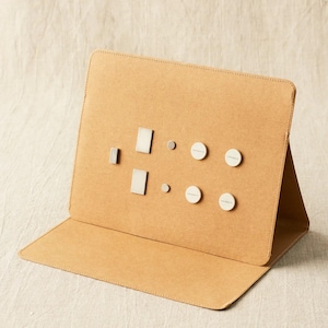 May include: A tan, foldable display board with a stitched border. It features several small, silver-colored rectangular and circular objects. The circular objects have white faces with text. The board is open, displaying the objects.