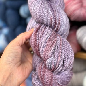 May include: A skein of lavender and burgundy yarn. The yarn is held in a hand and is in focus. Other skeins of yarn in various colors are visible in the background.