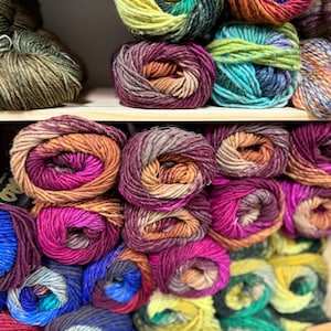 Op de afbeelding: Een plank toont diverse kleurrijke garenbollen. Het garen is in verschillende kleuren gerangschikt, waaronder groen, paars, oranje en blauw. Het garen lijkt zacht en geschikt voor brei- of haakprojecten. De bollen zijn strak opgerold.
