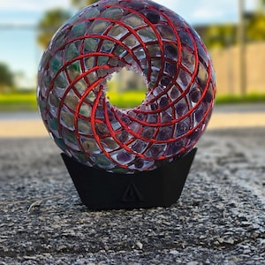 Può includere: Un'opera d'arte decorativa, sferica, con un'apertura centrale, composta da pezzi di vetro multicolore e filo rosso. La scultura poggia su una base nera angolare, mostrando un design unico.