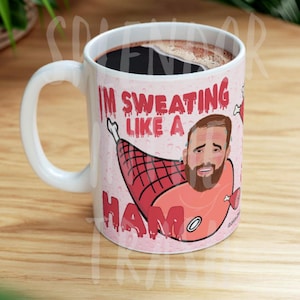 May include: White ceramic coffee mug with a humorous illustration of a man's face inside a ham, with the text "I'M SWEATING LIKE A HAM" in red. The mug is filled with dark coffee.