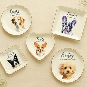 Op de afbeelding: Een verzameling gepersonaliseerde huisdierenschaaltjes in verschillende vormen, waaronder rond, vierkant en hartvormig. Elk schaaltje toont een aquarelportret van een hond, met de naam en data van het huisdier. De schaaltjes hebben een gouden rand.
