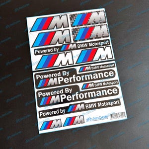 Op de afbeelding: Een vel met zelfklevende stickers met het BMW M-logo in blauw, rood en zilver, samen met de tekst Powered by M Performance en BMW Motosport. De stickers zijn in verschillende maten en stijlen gerangschikt op een wit achtergrondvel.