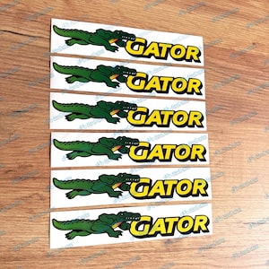 May include: Six rectangular white stickers arranged in a vertical column. Each sticker features a green alligator illustration on the left and the word GATOR in bold yellow capital letters with a black outline on the right side of the design.