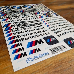 May include: A white sticker sheet featuring various BMW Motorsport and M Performance logos in black, blue, and red. The sheet includes multiple text-based decals, circular emblems, and carbon-fiber patterned squares with the M logo and a barcode.