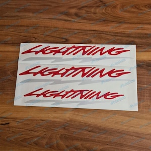 May include: A set of three adhesive decals on a white backing sheet. Each decal features the word LIGHTNING in a bold, red, stylised script font, positioned above a light grey, jagged, horizontal lightning bolt graphic element.