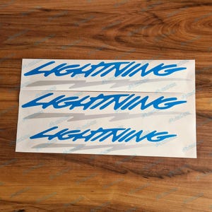 Op de afbeelding: Een set van drie witte rechthoekige stickers met het woord Lightning in een vet blauw scriptlettertype. Elke sticker bevat een gestileerde lichtgrijze bliksemschichtgrafiek die direct onder de blauwe tekst is geplaatst.