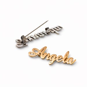 May include: Two name pins are shown on a white background. One is silver and reads "Samantha" in cursive. The other is gold and reads "Angela" in cursive. Both have pin backings for attachment.