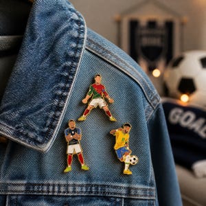 以下が含まれることがあります： デニムジャケットに飾られた、3つのサッカー選手のエナメルピン。ピンは、サッカーボールを蹴る選手など、アクションポーズの選手を特徴としています。ピンは、ポルトガル、フランス、ブラジルの代表チームの色です。